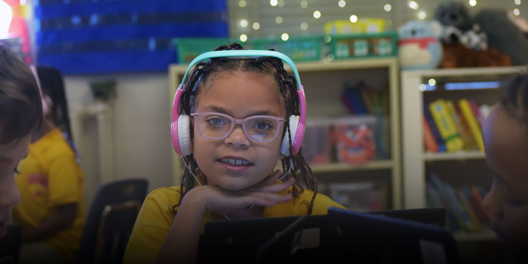 Student in pink headphones and glasses using a laptop in a classroom.